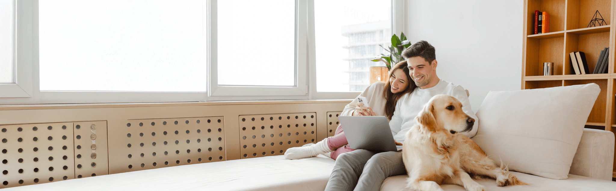 A man and woman smiling and sharing a computer on the couch. They are sitting next to a dog and working together on their PC.