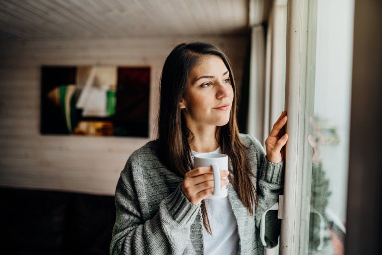 A woman drinking coffee and looking out of a porch window.